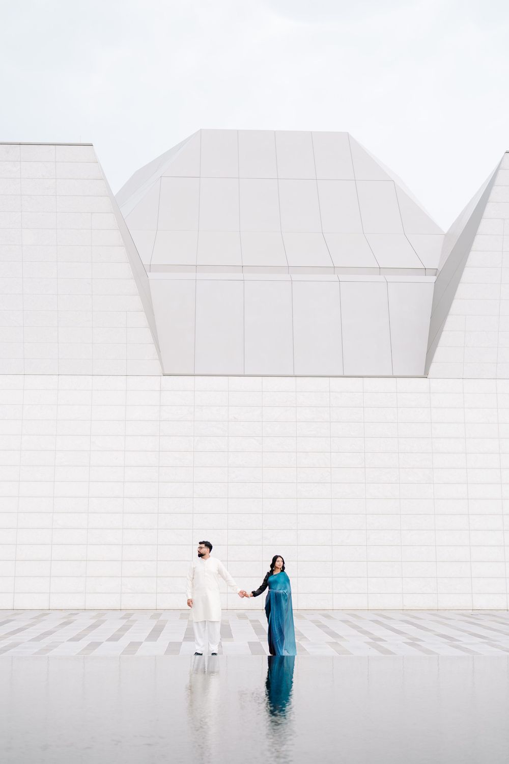 Couple in traditional South Asian attire holding hands near a modern white geometric building with reflective water.