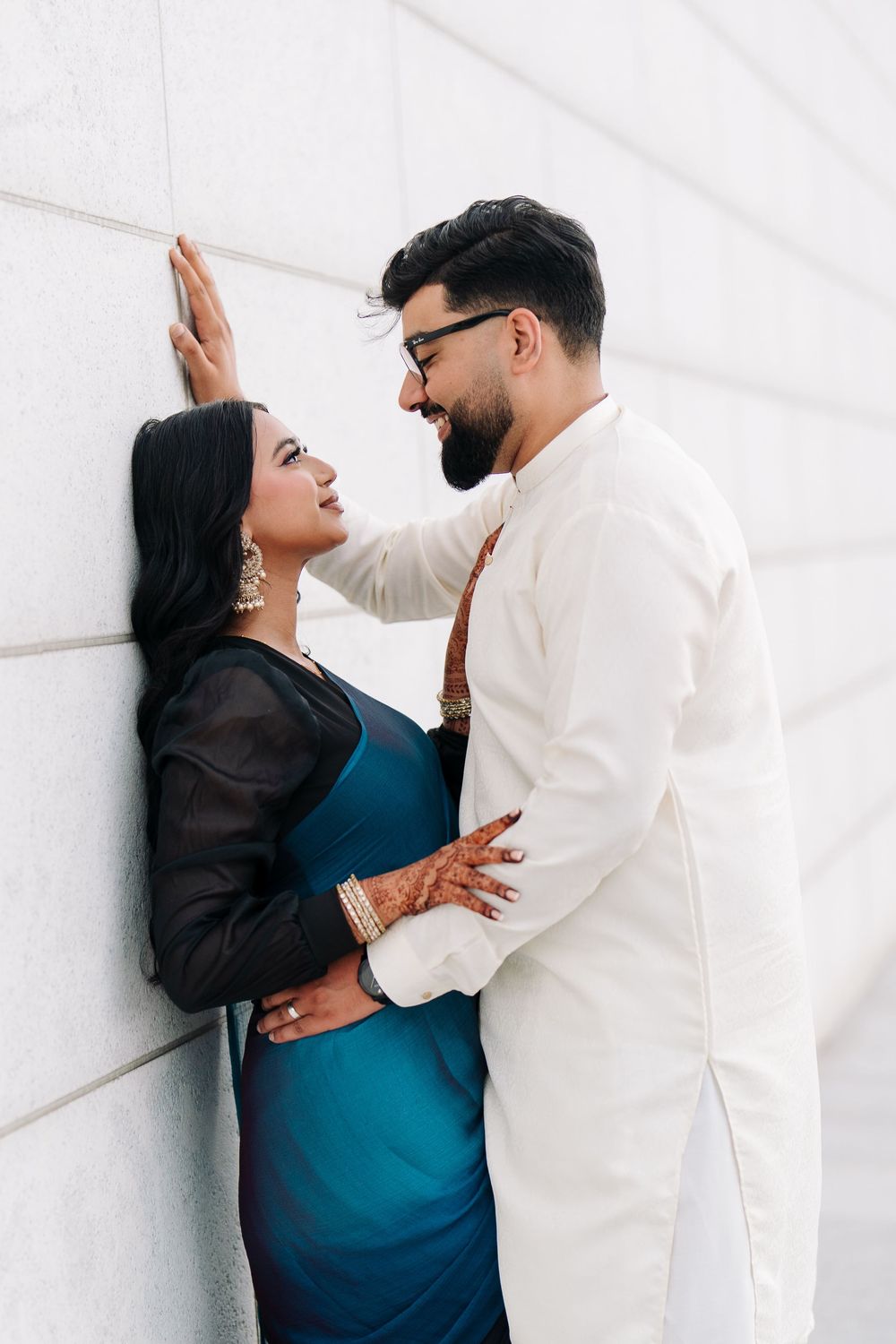Indian couple in traditional attire sharing a romantic moment against a white wall, woman in teal saree, man in white kurta.