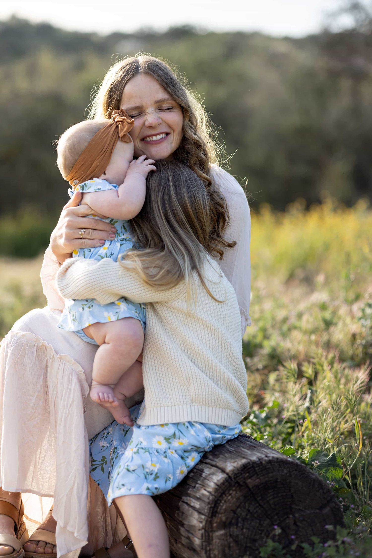 Smiling mother hugging baby outdoors on a sunny day, sitting on a log in a green field.