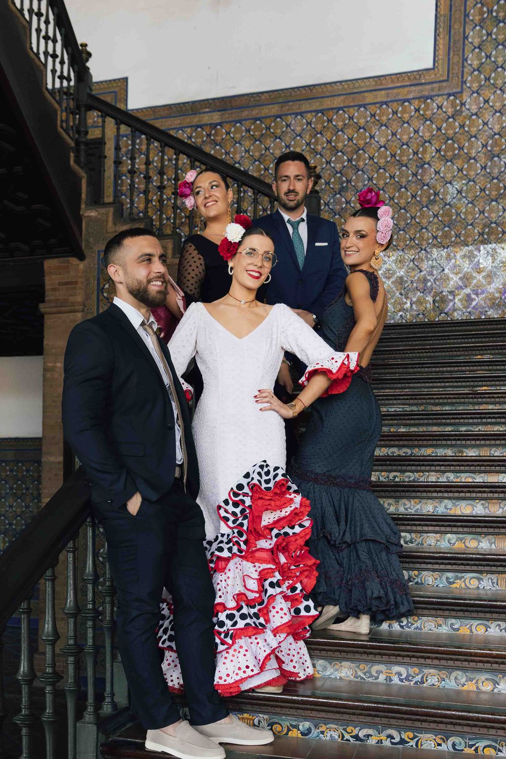 Group of five people in flamenco attire posing on ornate tiled staircase, women in ruffled dresses with flowers in hair.