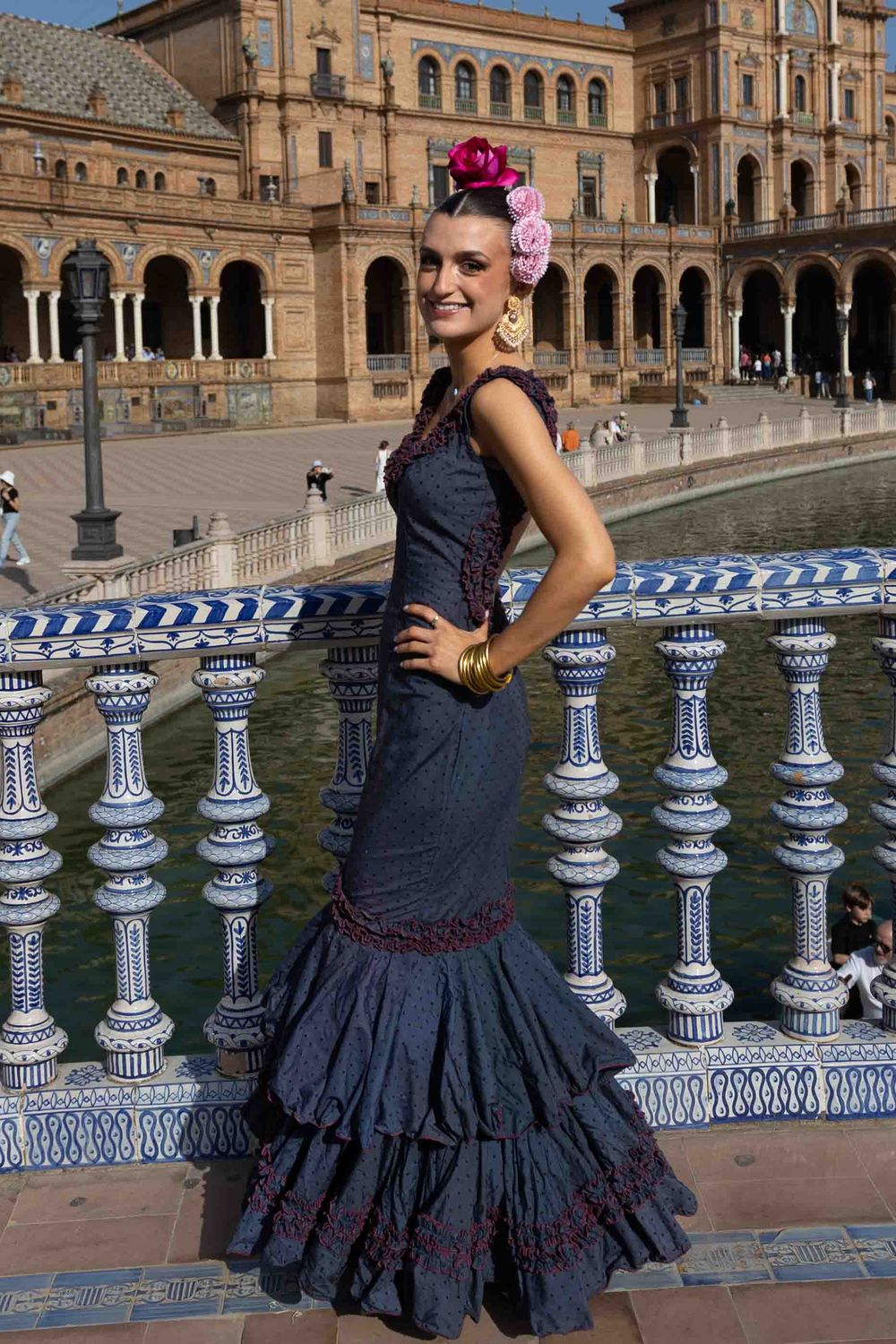 A smiling woman in a traditional flamenco dress with flowers in her hair poses at Plaza de España, Seville, Spain.