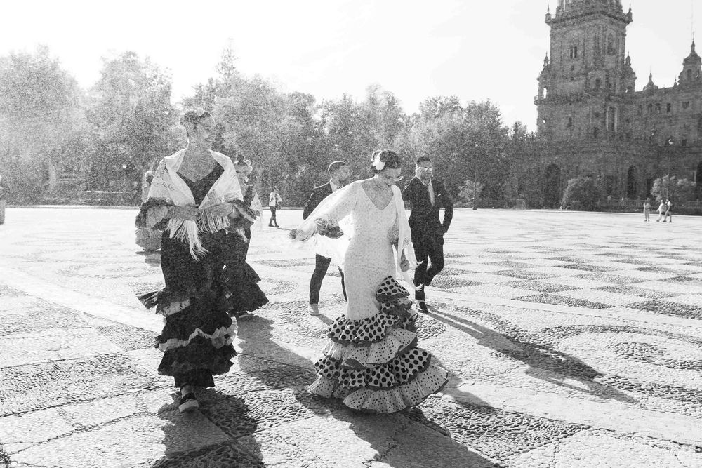 Two women in traditional flamenco dresses walk across a sunlit plaza in Seville, Spain.