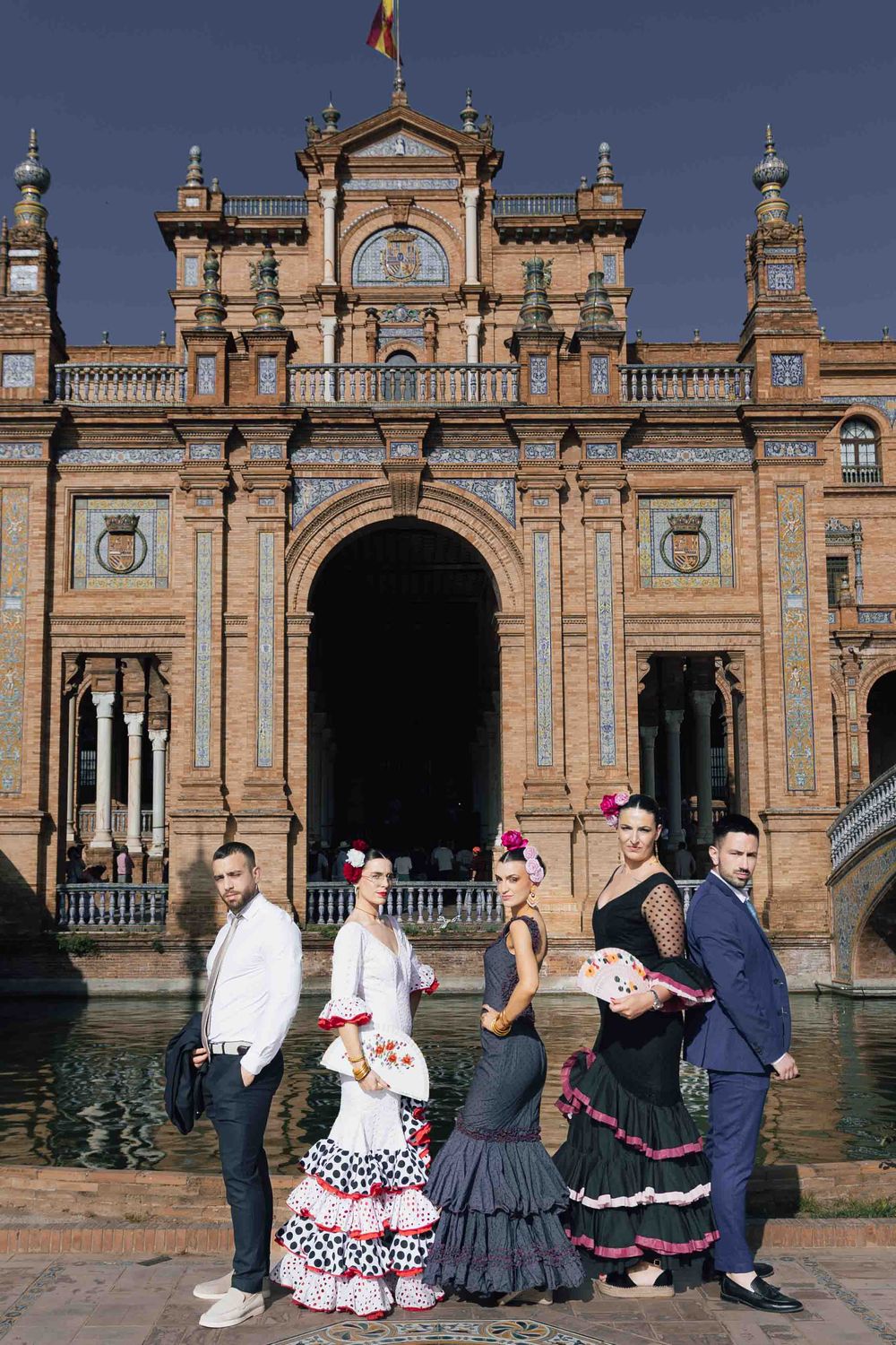 Group of flamenco dancers pose in traditional attire before Plaza de España's ornate facade in Seville.