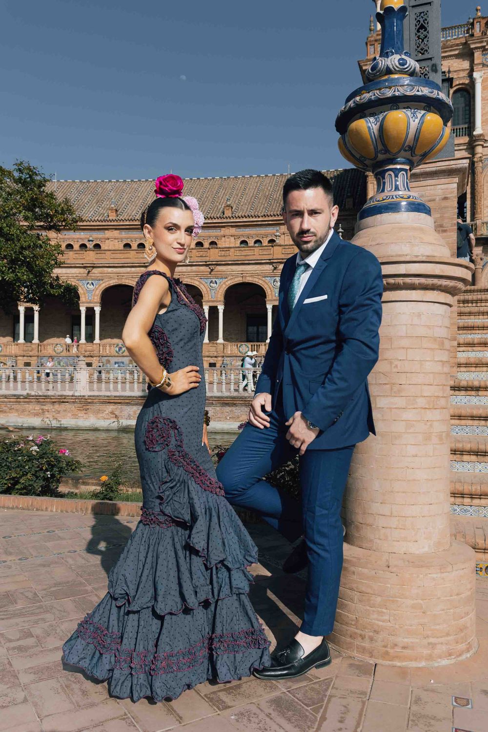 Flamenco couple posing at Plaza de España Seville, woman in ruffled dress with flower, man in blue suit.