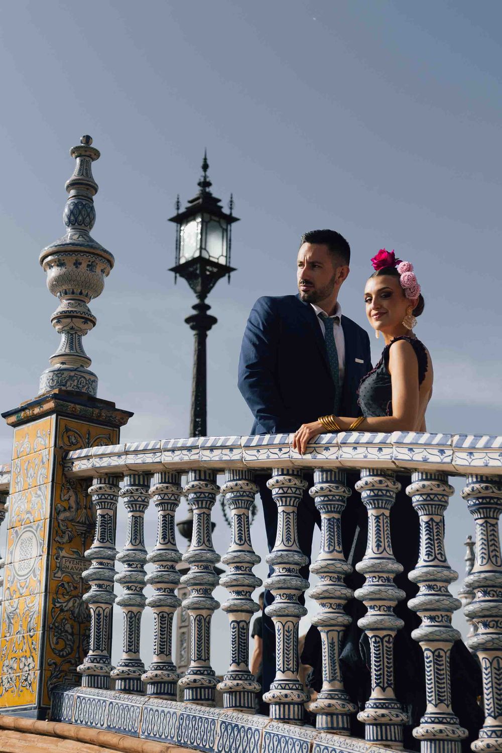 Elegant couple in formal attire stand on ornate tiled balustrade at Plaza de España, Seville.
