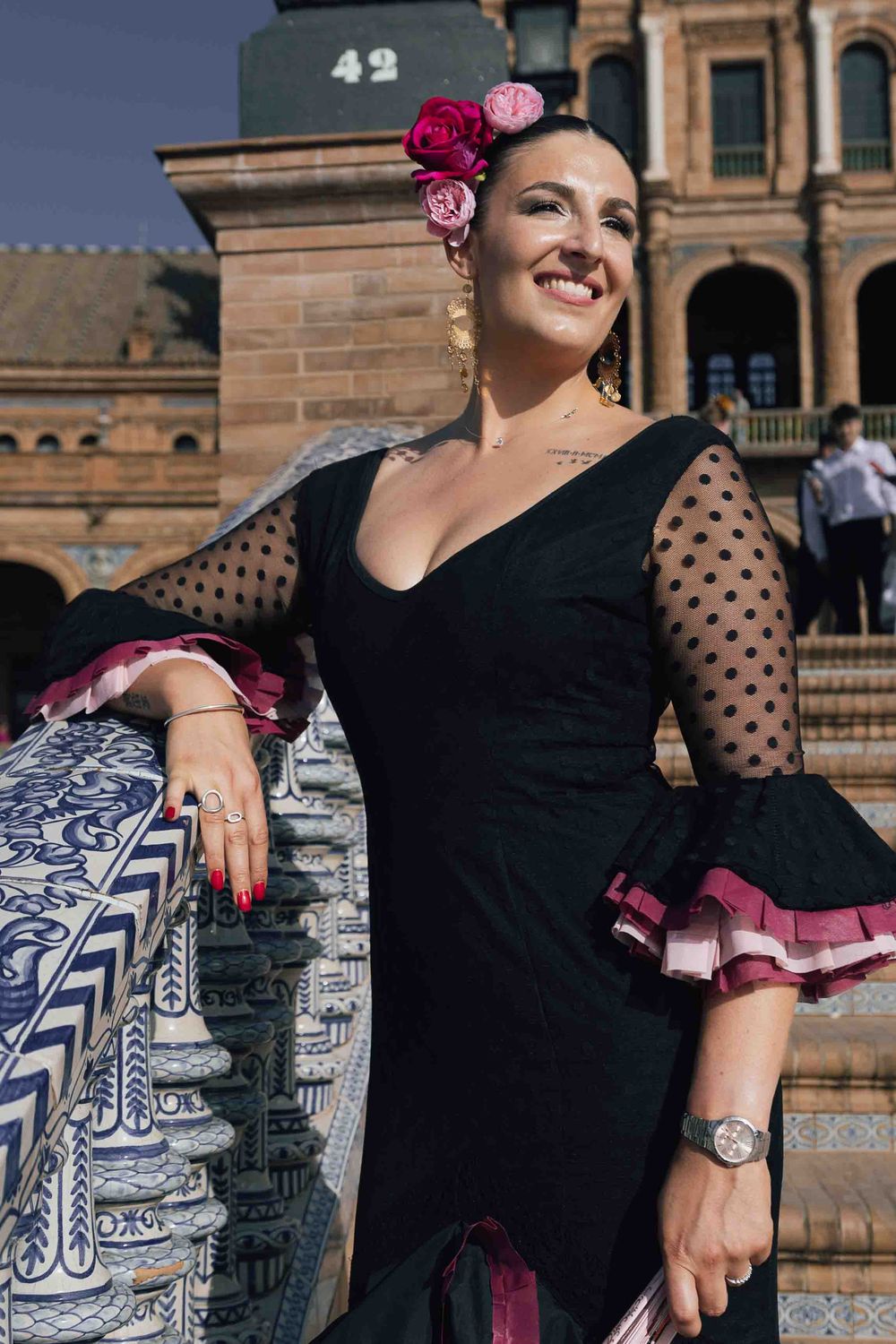 Woman in black flamenco dress with polka dot sleeves and pink flowers, posing at Plaza de España, Seville.