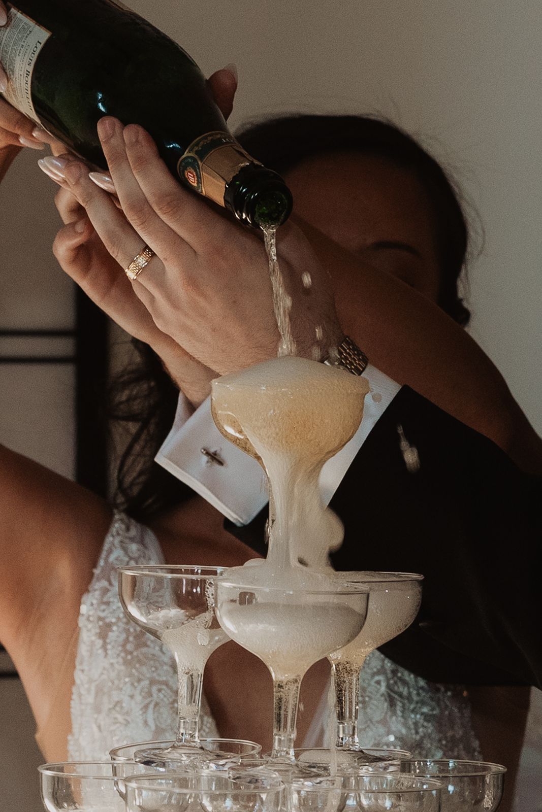 Bride pouring champagne into a tower of coupe glasses at a wedding reception celebration.