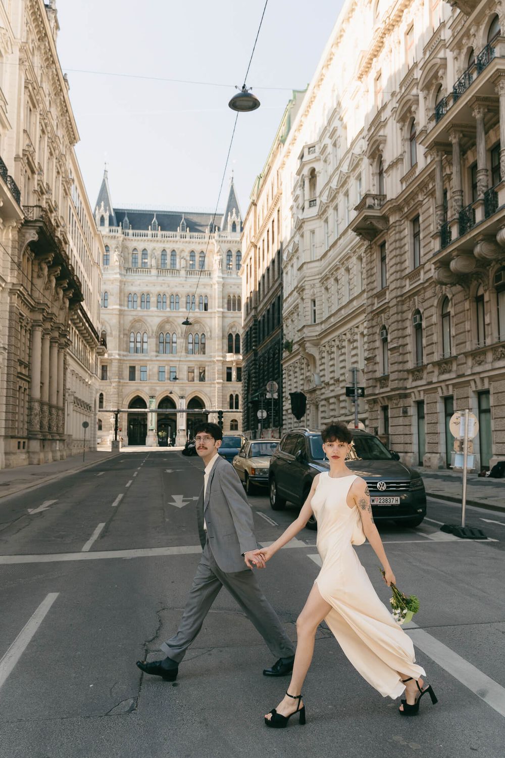 Bride and groom holding hands walking across a street in Vienna, Austria, with historic European architecture in the background.