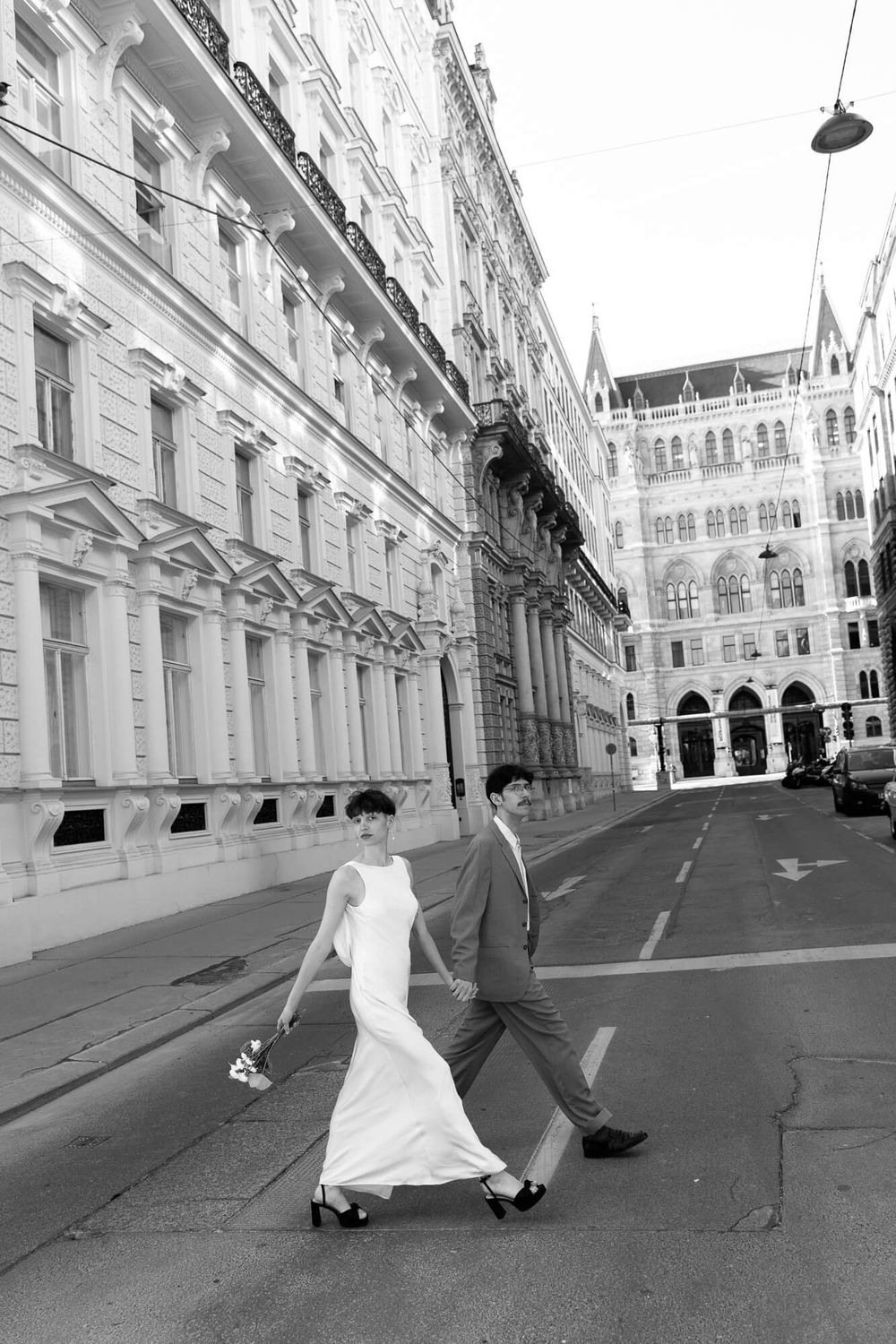 Black and white photo of a bride and groom walking hand in hand on a city street with grand European architecture.