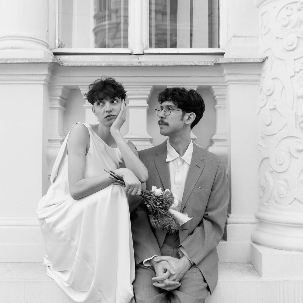 Black and white photo of a young couple posing elegantly, woman in white dress holding flowers near a classic building.