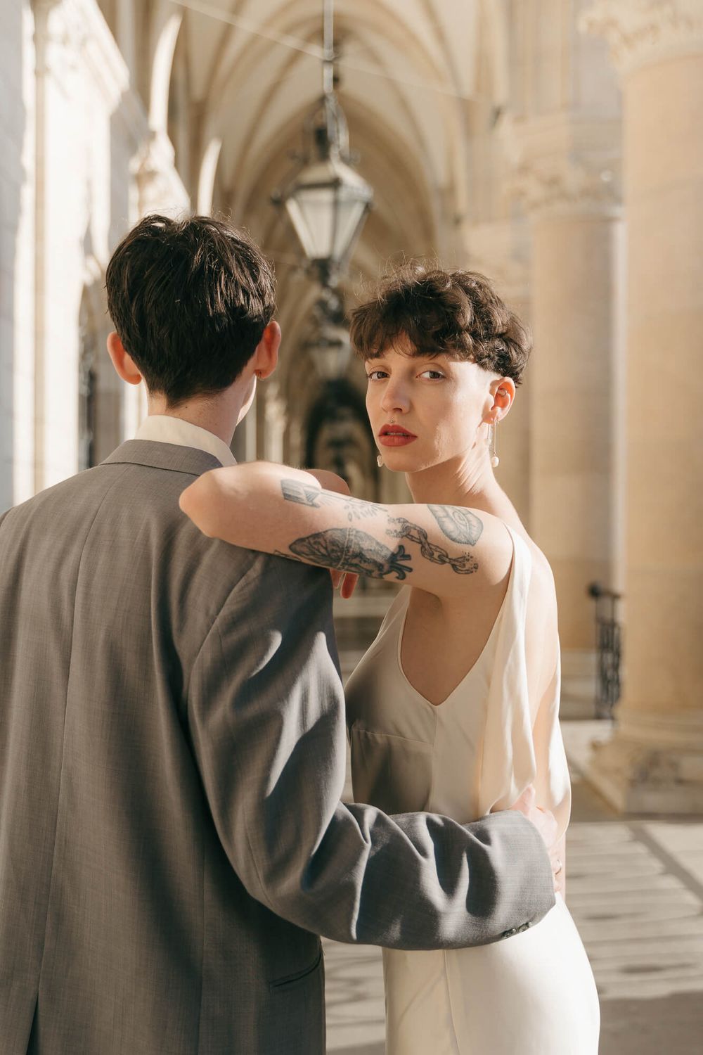 Tattooed bride in white satin dress embraces groom in gray suit under gothic stone arches.