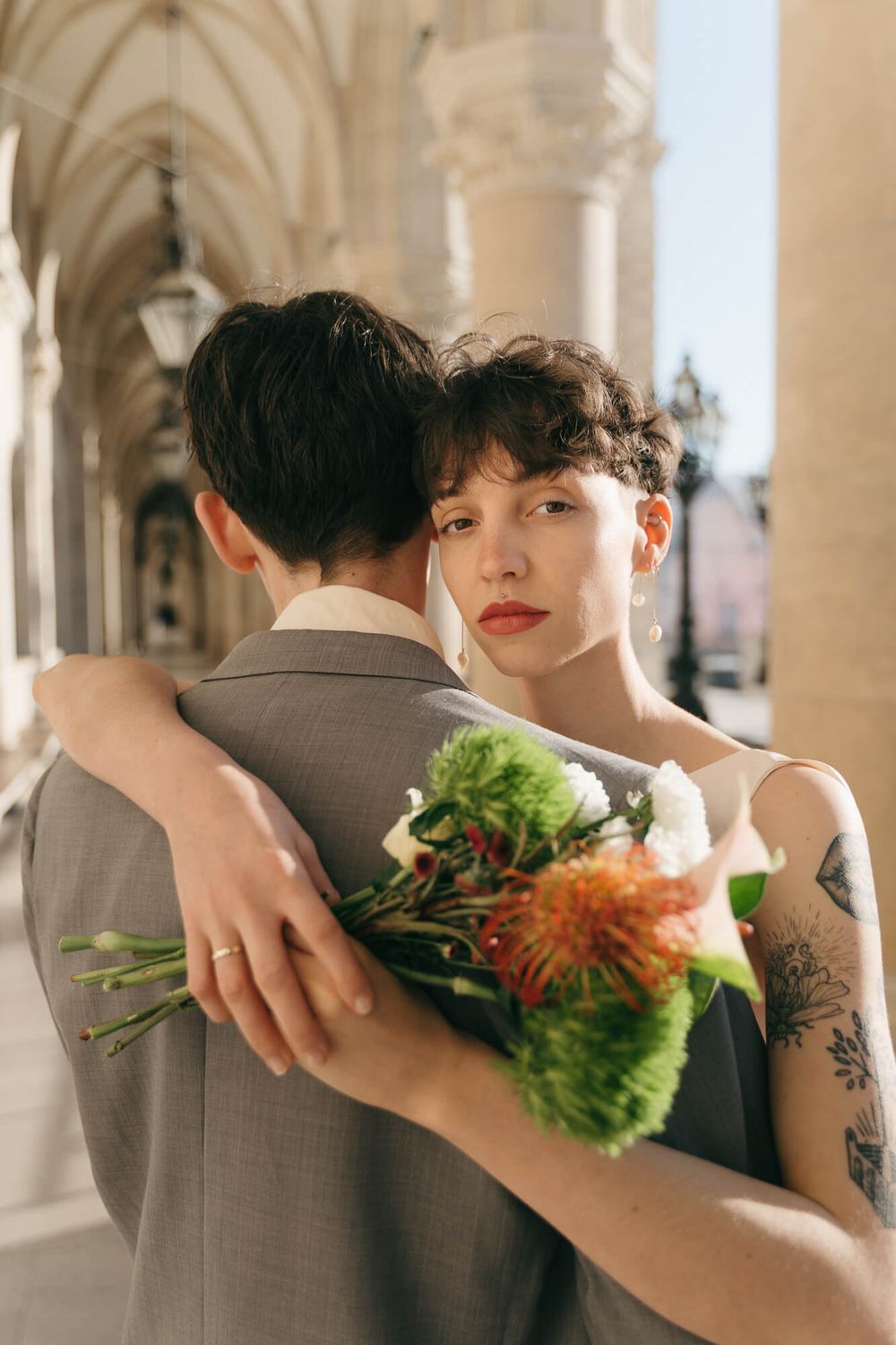 Bride with tattoos hugs groom holding colorful wedding bouquet under elegant stone archway.
