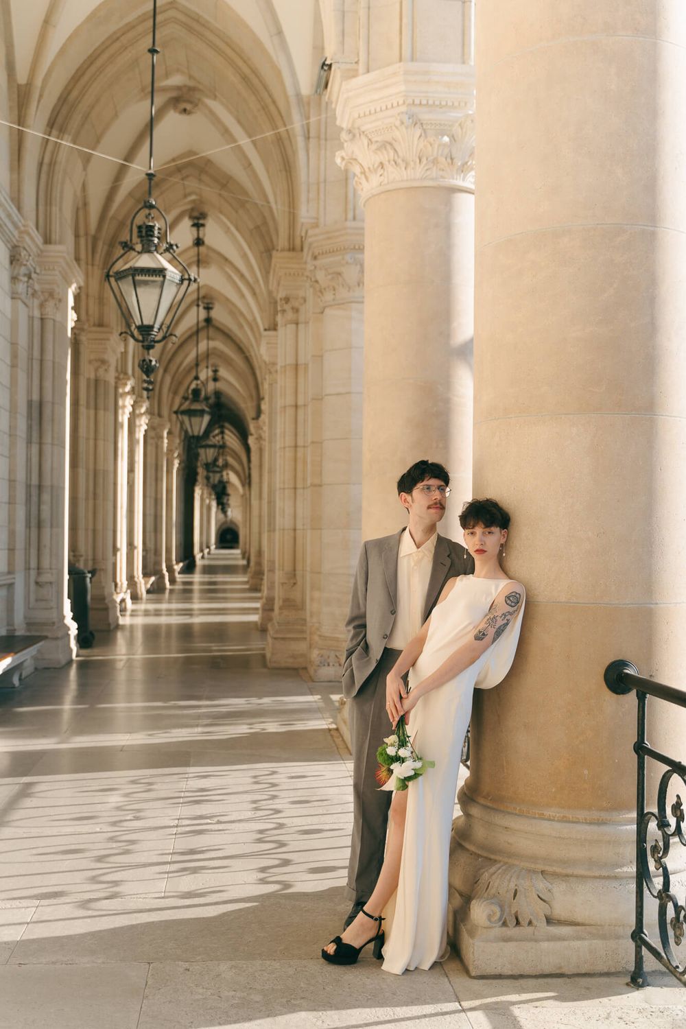 Elegant couple in wedding attire pose beside ornate stone columns in a grand gothic corridor.