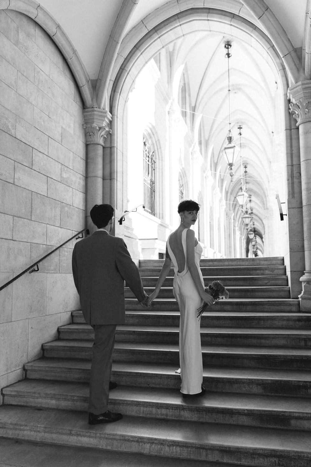 Black and white photo of a bride and groom holding hands walking up stone steps under gothic arches.