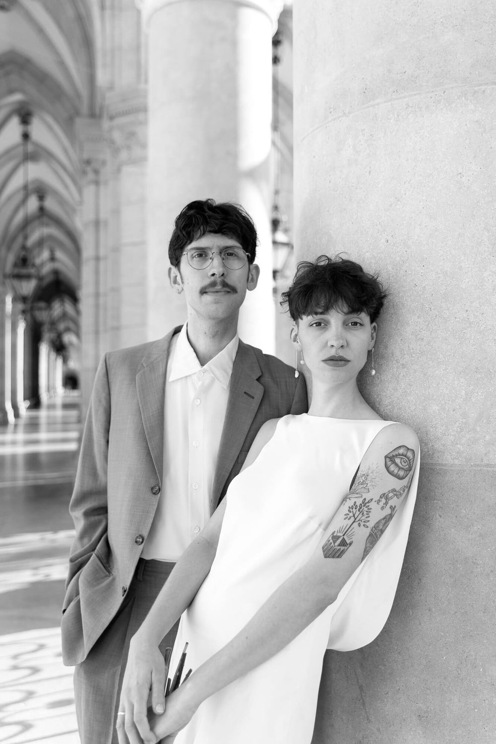 Black and white portrait of a couple in wedding attire posing by stone columns in an elegant archway.