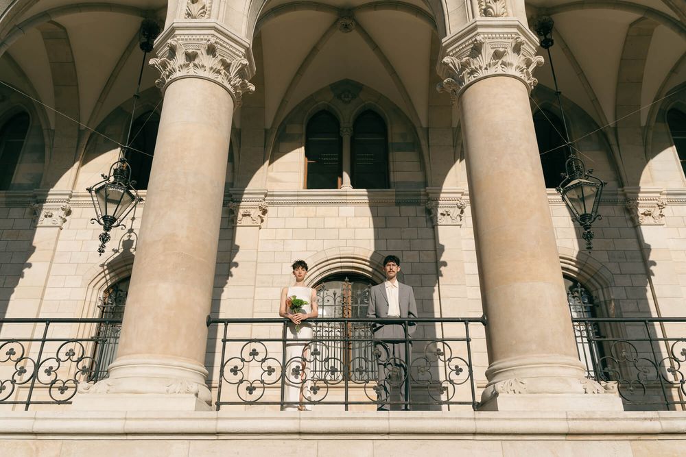 A wedding couple stands on a balcony beneath grand Gothic stone arches of a historic building.