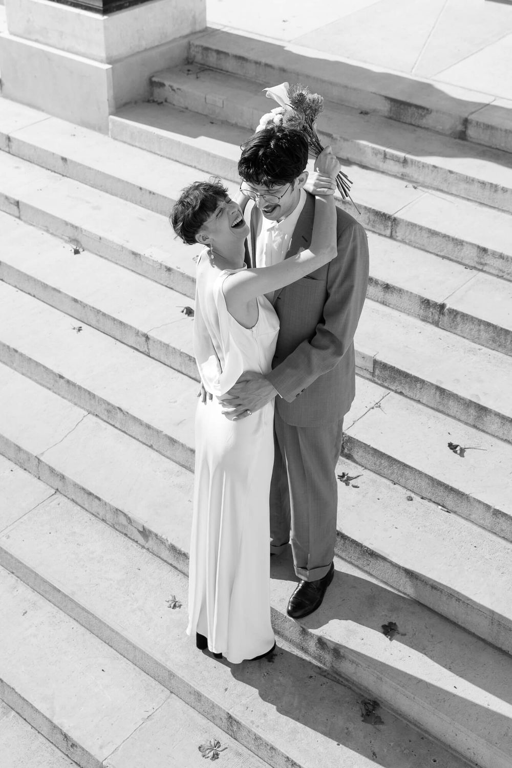 Black and white photo of a bride and groom embracing on stone steps, she holds a bouquet while they gaze at each other.