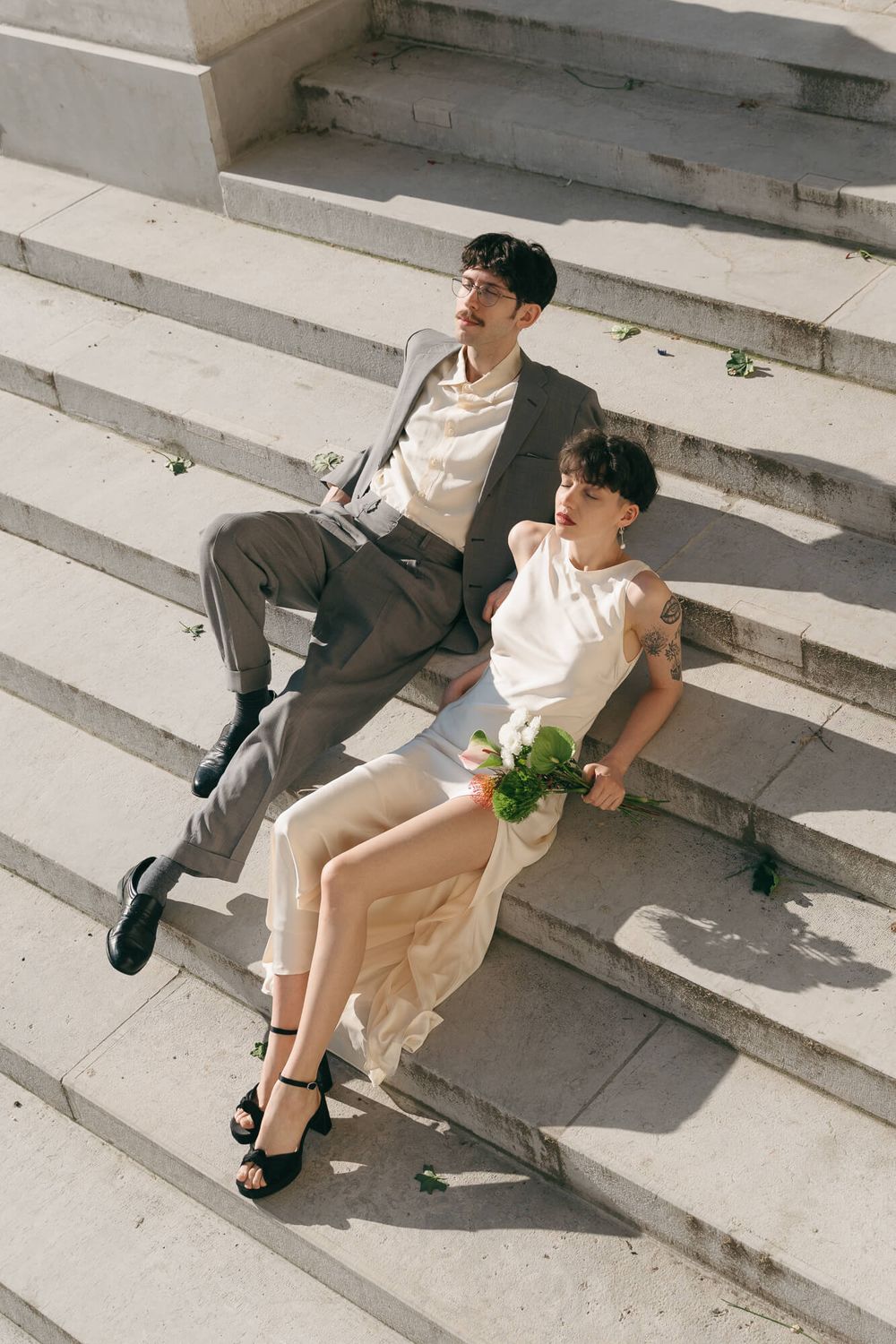 A bride and groom sit on sunlit stone steps, she holds a white bouquet while both look away in opposite directions.