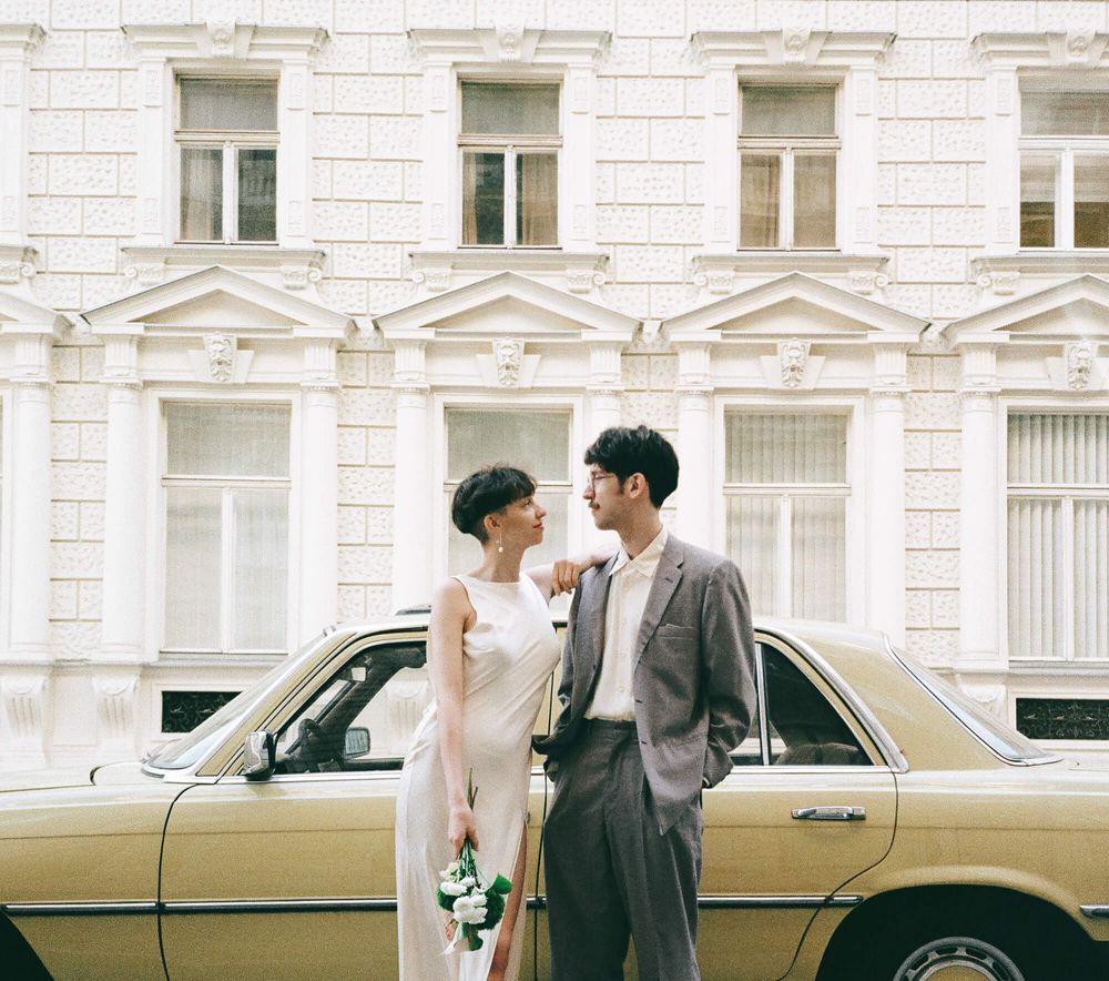A couple in wedding attire stands beside a vintage yellow car in front of a classic European building.