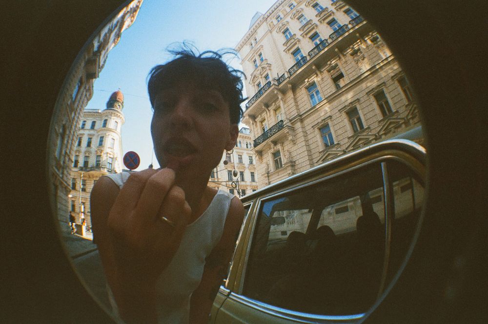 Young man photographed with fisheye lens on a sunny European city street beside a vintage yellow car.