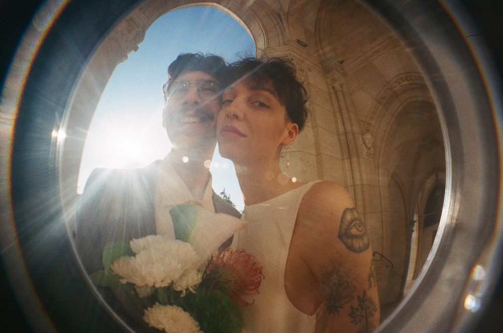 Couple posing for a wedding photo through a circular lens with sunlight and white bouquet.