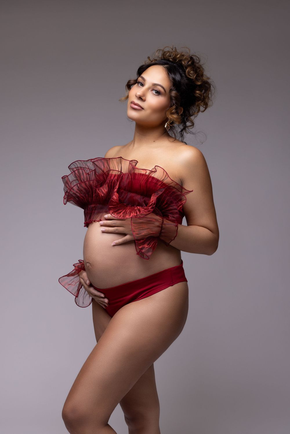 Maternity portrait with red tulle wrap and matching bottoms against grey studio backdrop.