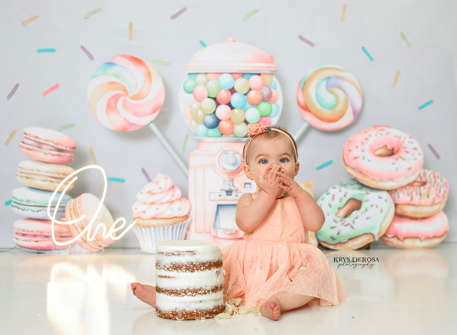 Baby girl in peach dress sits before birthday cake at candy-themed first birthday photo shoot with donuts and lollipops.