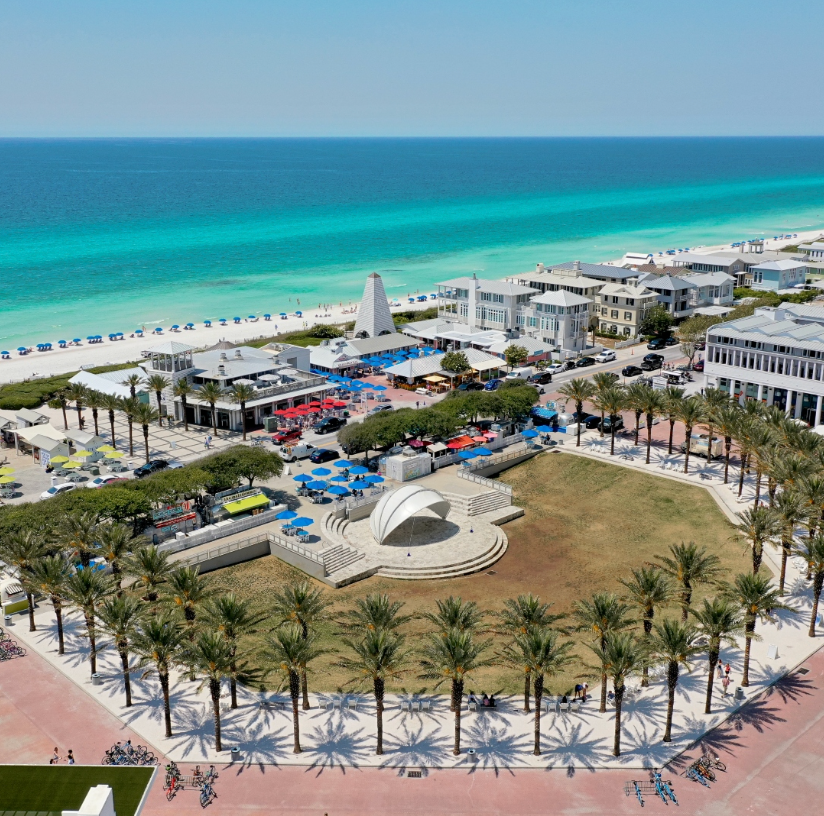 Aerial view of a beachfront town with turquoise Gulf waters, palm trees, and a central park plaza in Seaside, Florida.
