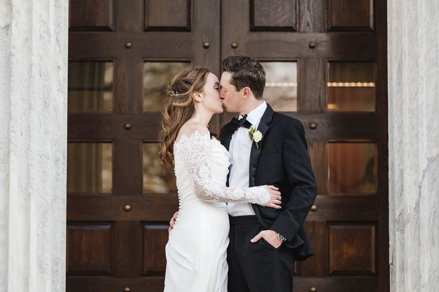 Bride and groom share a romantic kiss before grand wooden doors flanked by white marble columns.