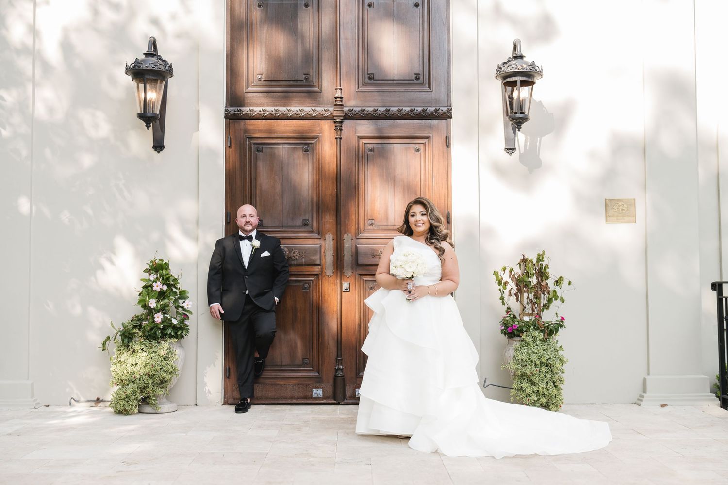 Bride and groom pose elegantly before ornate wooden doors with lanterns and lush greenery.