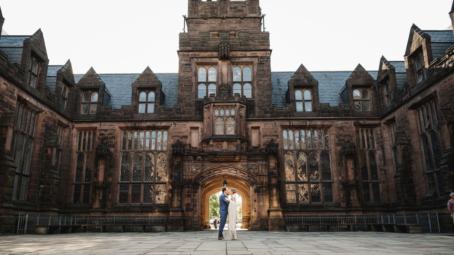 Couple shares a romantic moment under a grand Gothic stone archway at a historic university building.