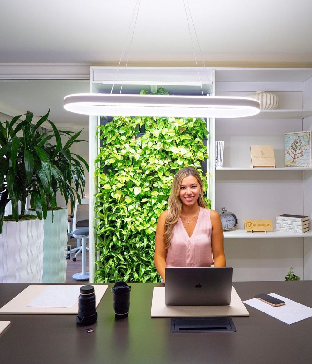 Woman smiling at desk with laptop in modern office featuring green living wall and oval pendant light.