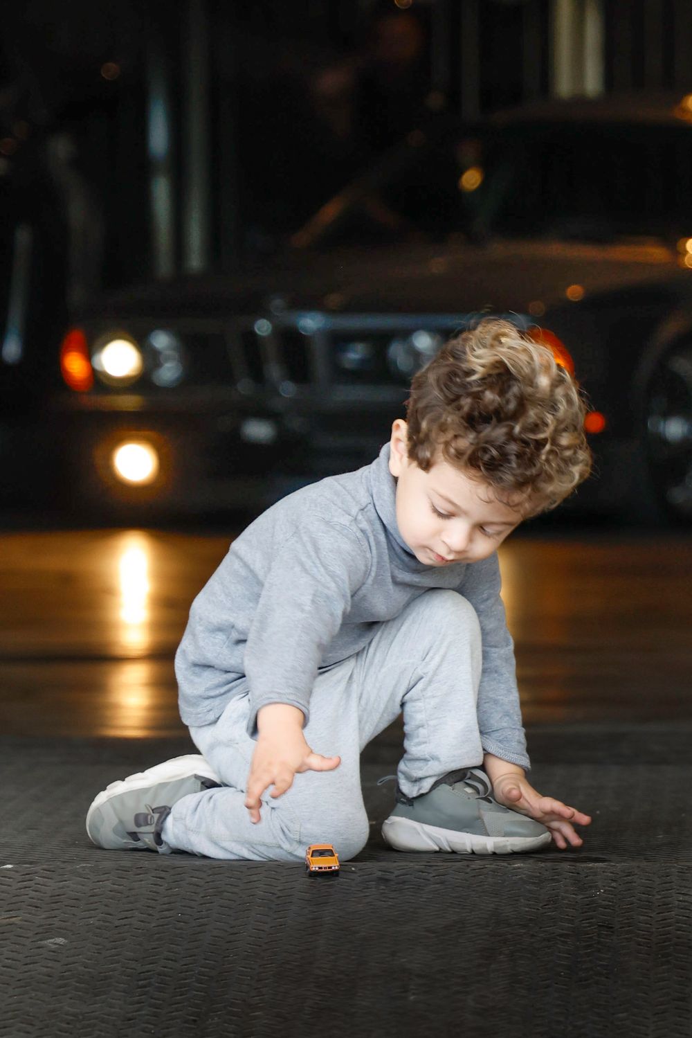 Young curly-haired boy in gray outfit playing with a small toy car on the floor near a parked vehicle.