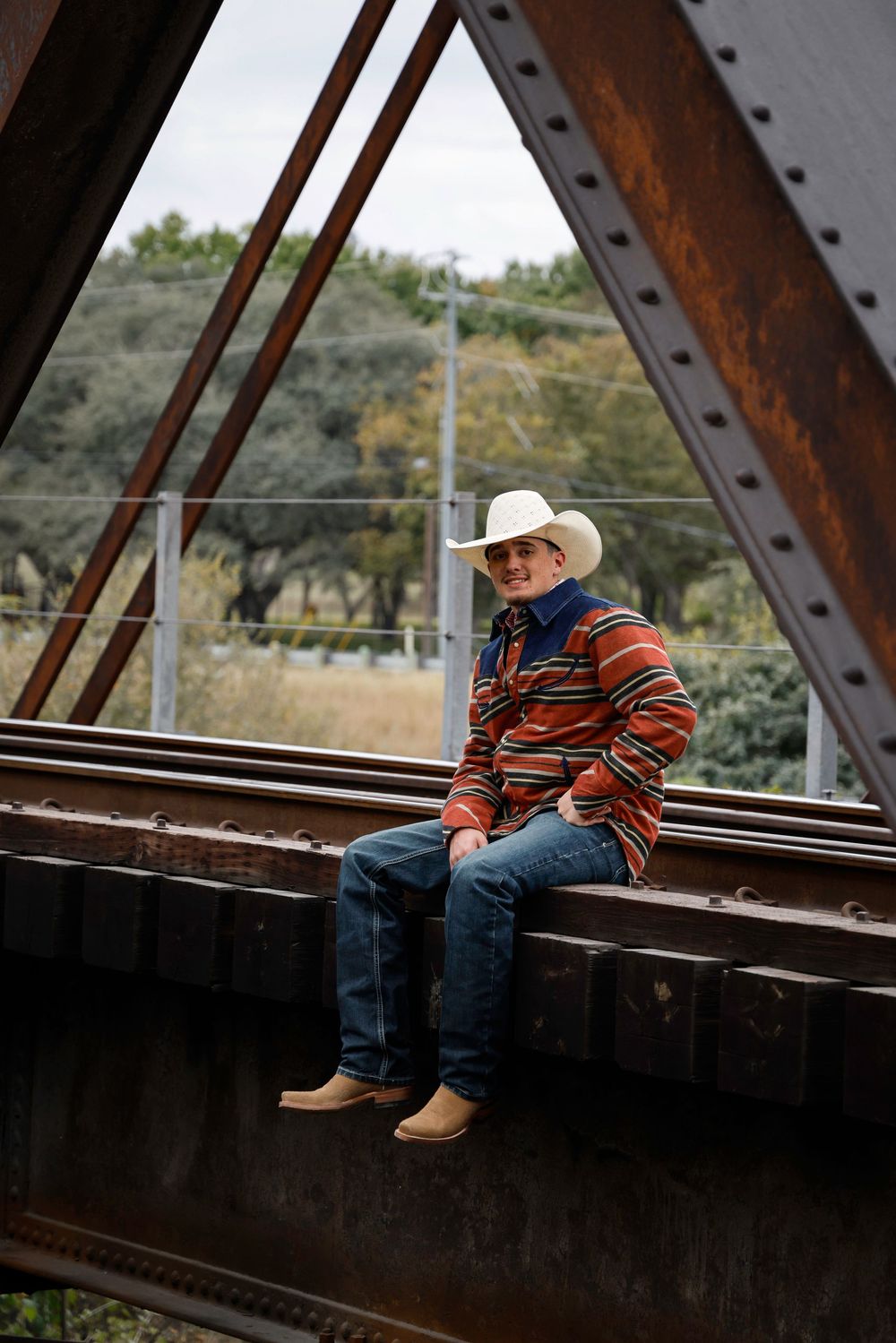 A man in a white cowboy hat, striped jacket, jeans, and boots sits on a rustic iron bridge outdoors.