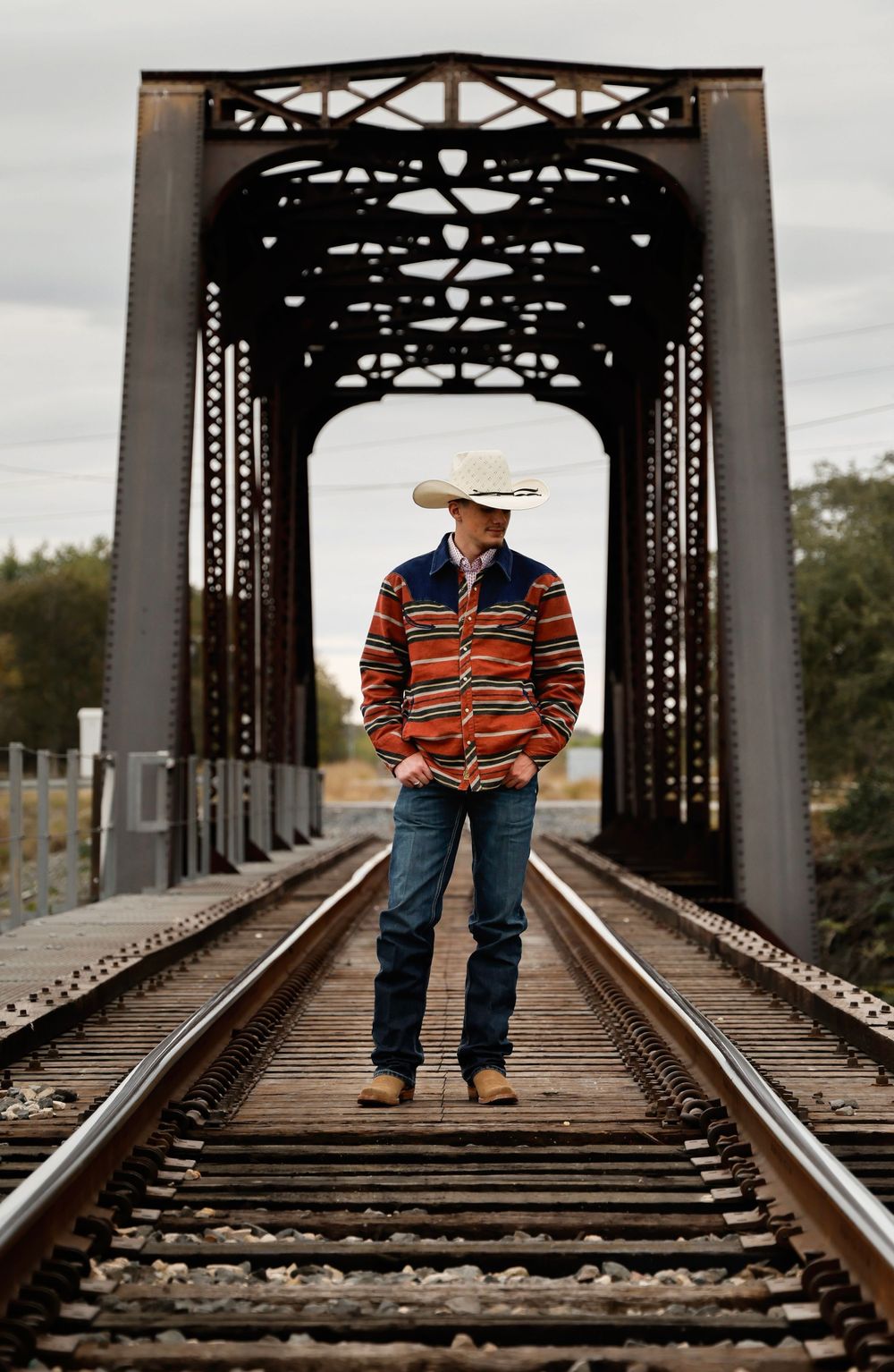 Man in cowboy hat and striped jacket standing on railroad tracks beneath a steel bridge.
