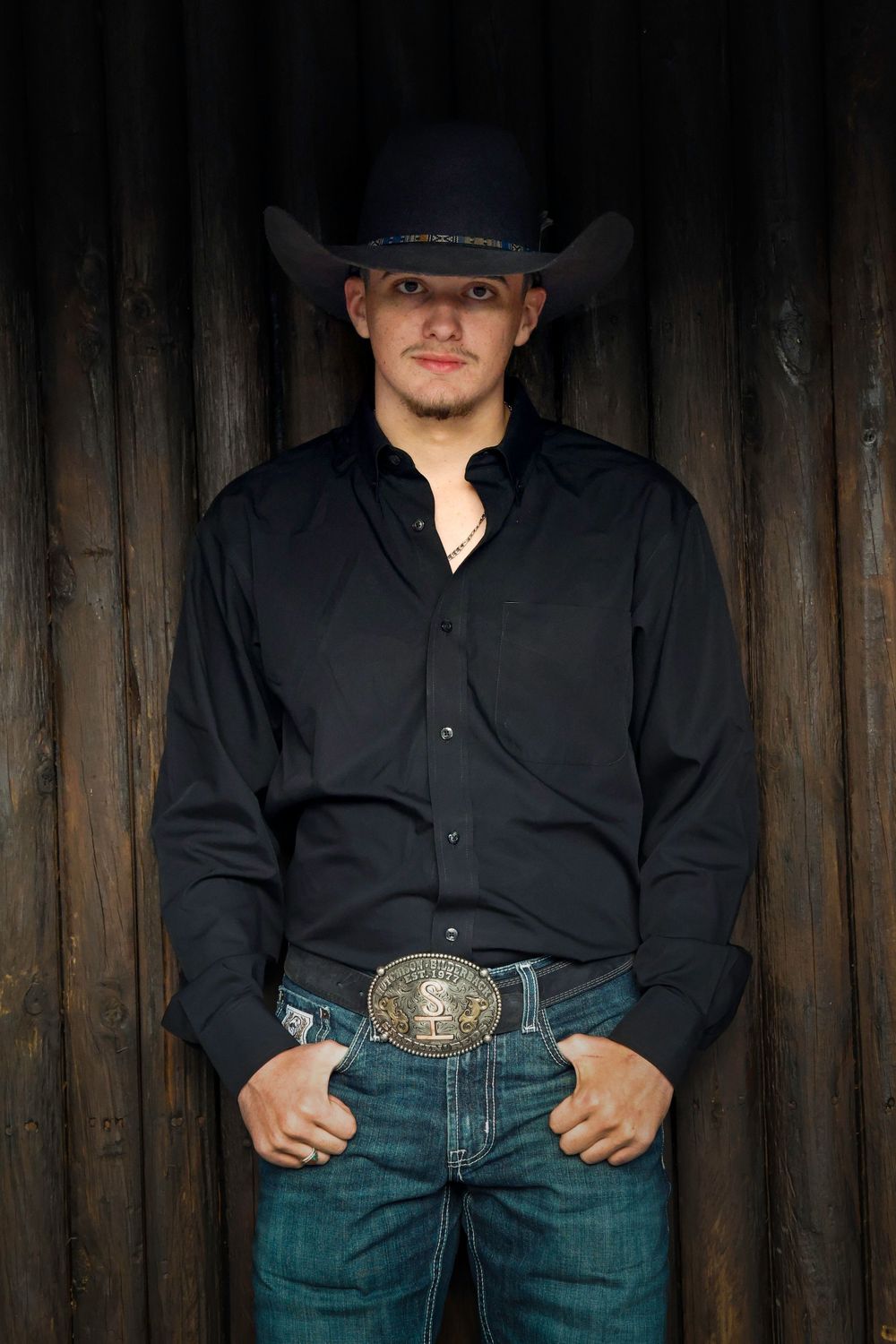 Young man in black cowboy hat, black western shirt, jeans, and decorative belt buckle posing against rustic wood wall.