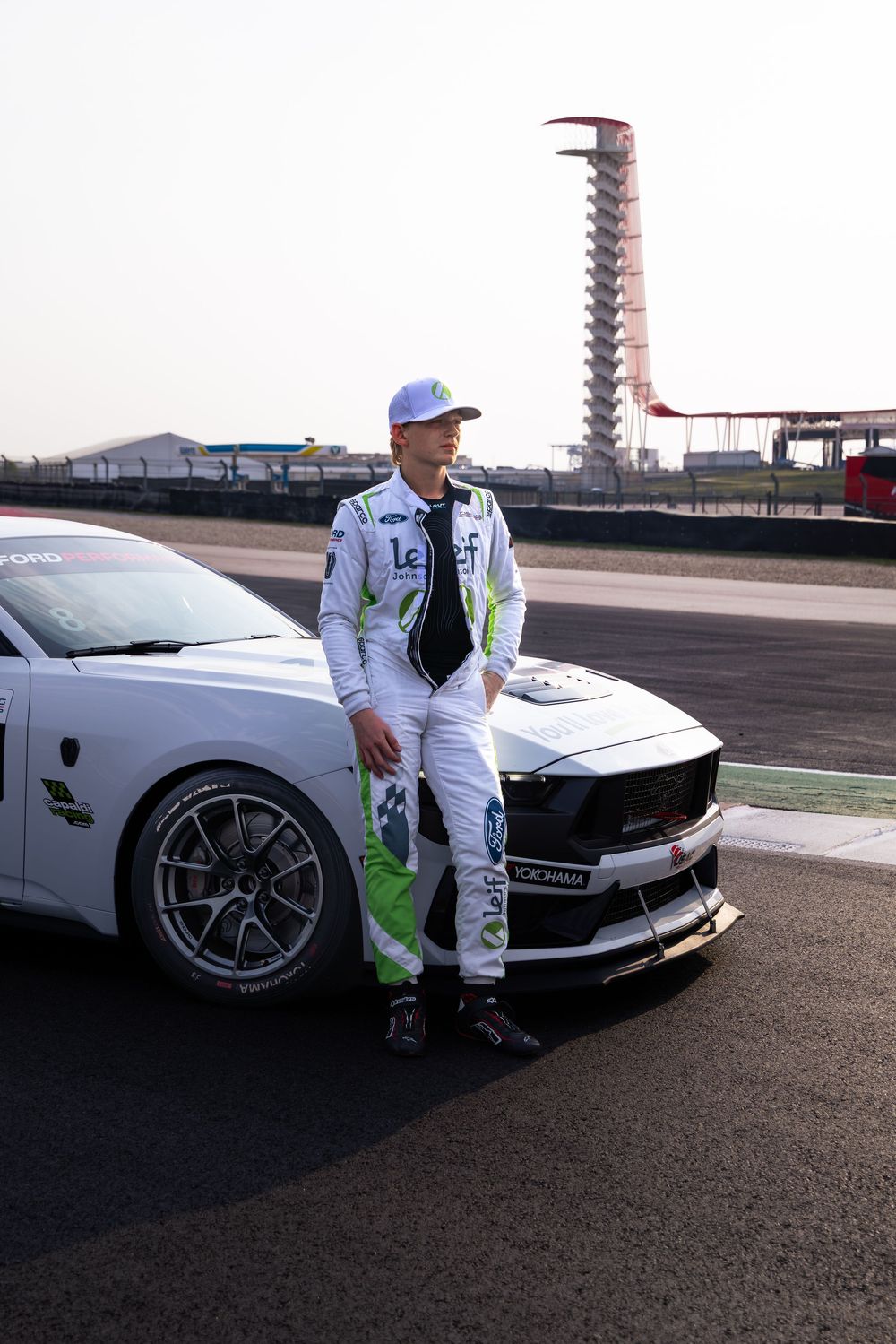 Race car driver in white and green suit leaning against a white Ford Mustang GT race car on a professional circuit track.