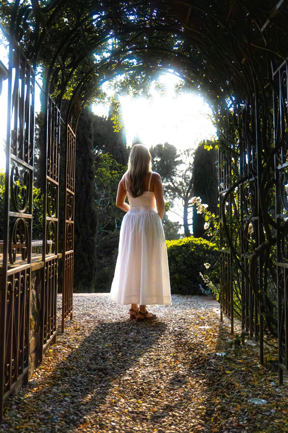 A woman in a white dress stands at the end of a vine-covered iron archway bathed in sunlight.