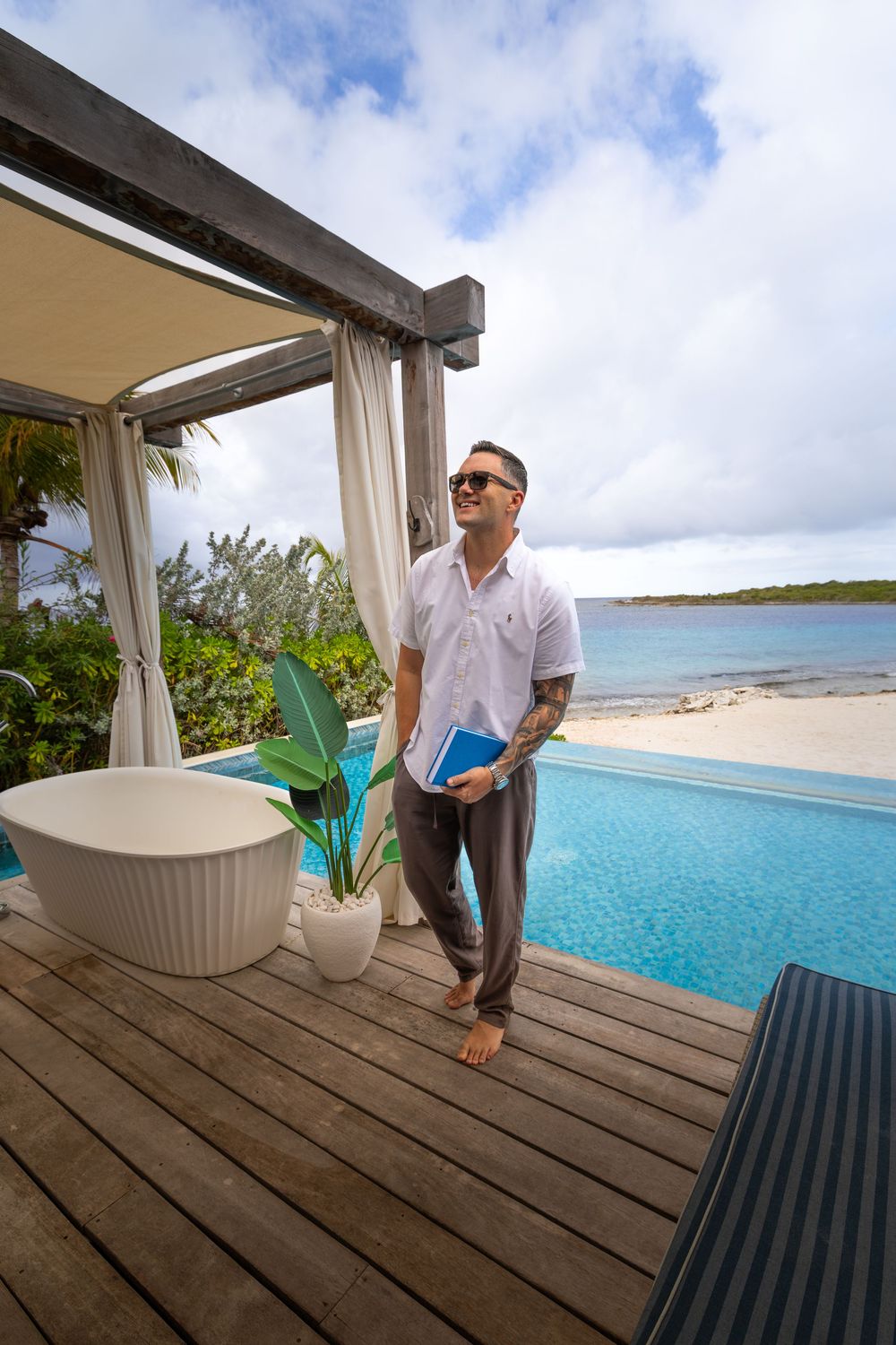A tattooed man in sunglasses stands by an infinity pool overlooking a tropical beach resort.