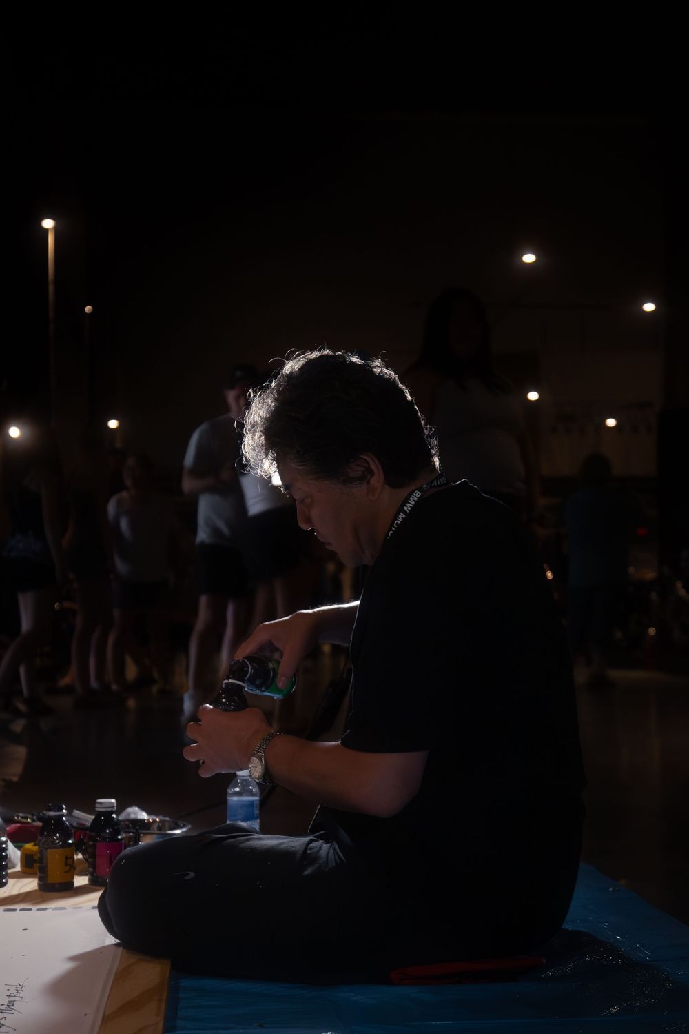 Person sitting cross-legged on floor at night, working with tools and supplies spread out, with people standing in background.