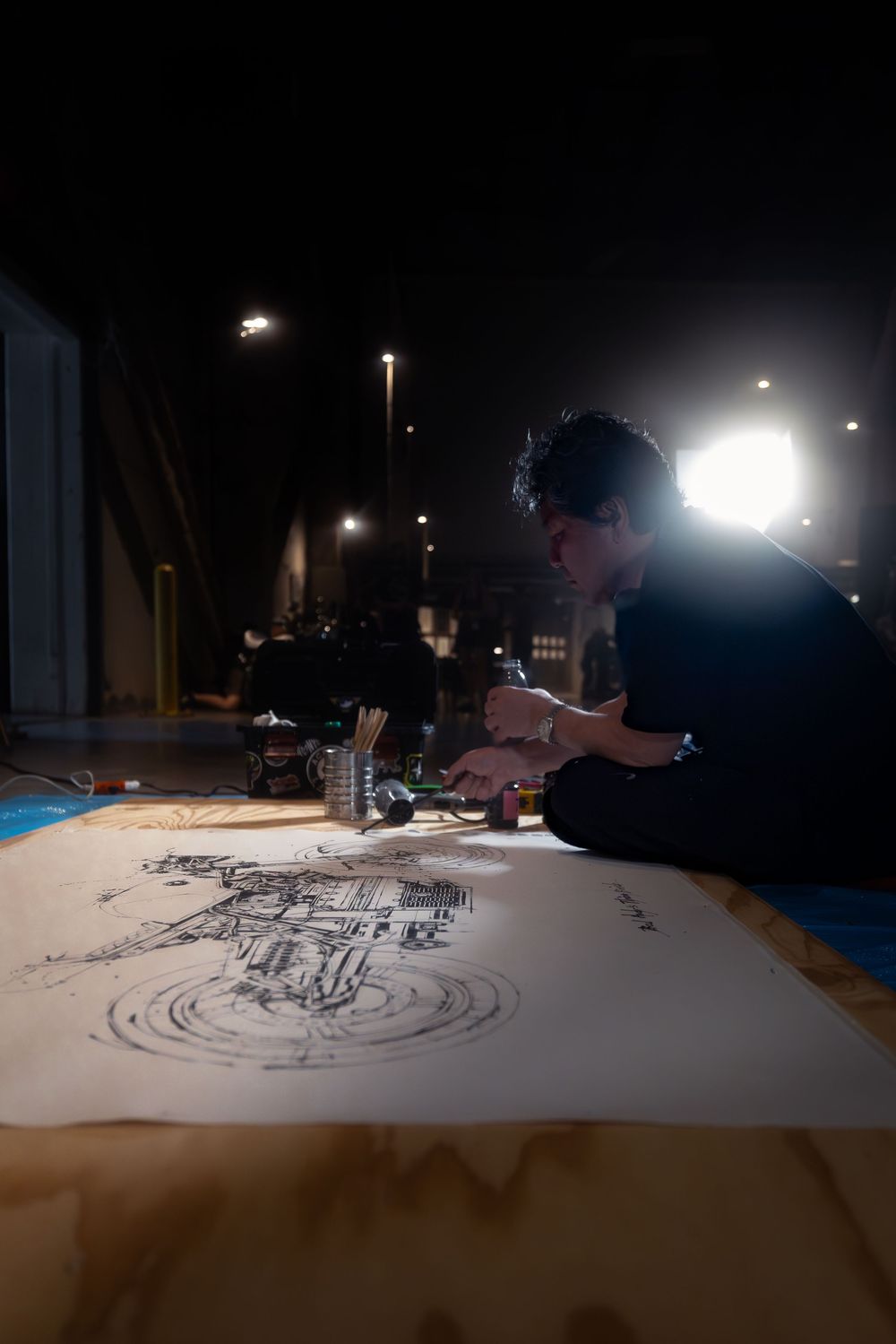 Artist sitting on floor drawing detailed architectural sketch on large paper, illuminated by bright studio light in dark room.