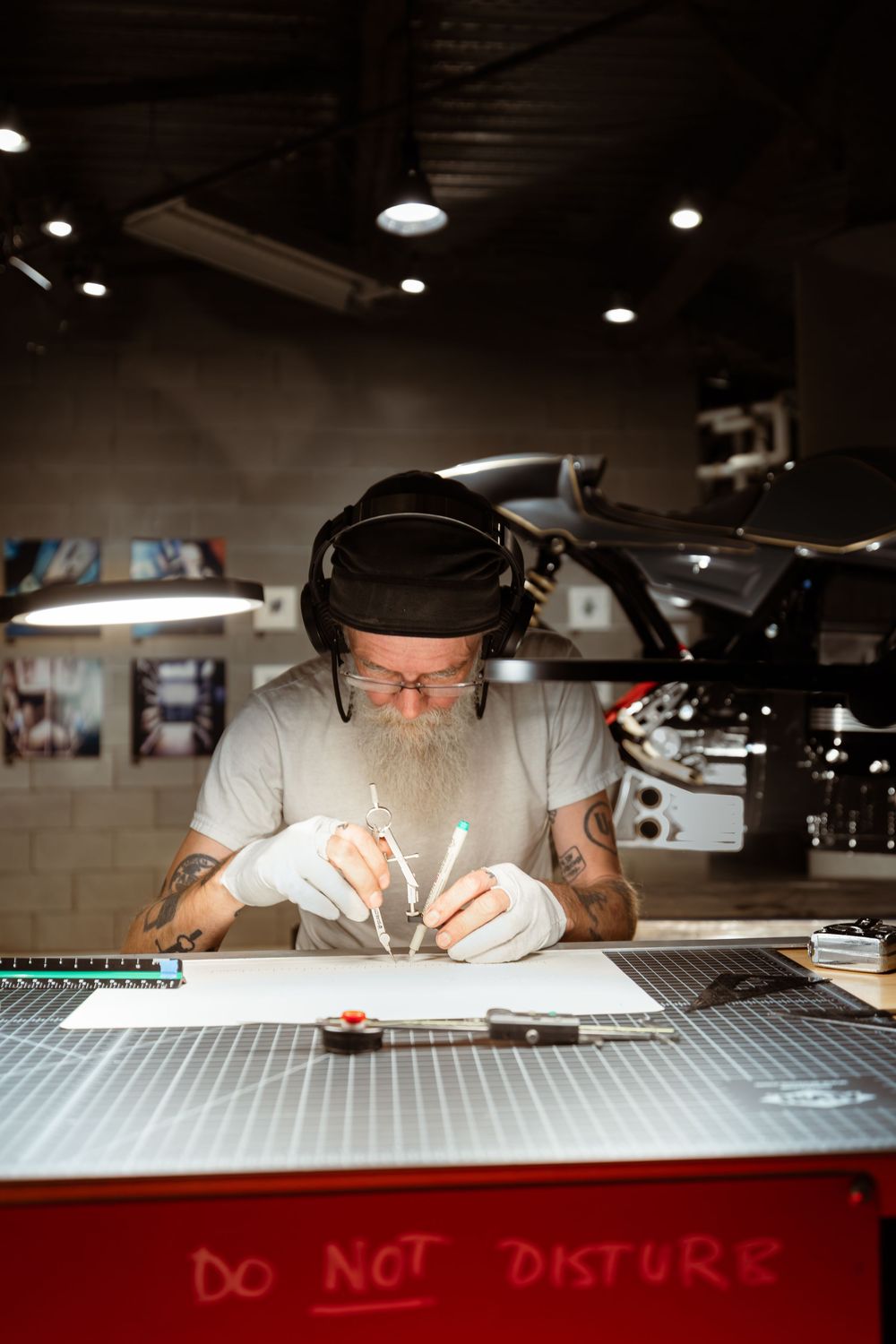 A bearded tattooed man works carefully at a drafting table in a garage workshop.