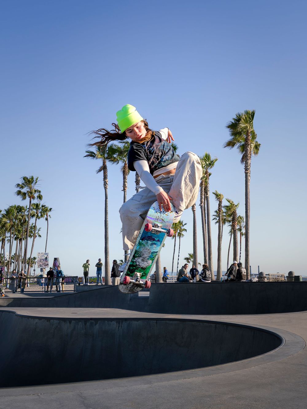 Skateboarder performing a trick at Venice Beach skate park with palm trees and blue sky in the background.