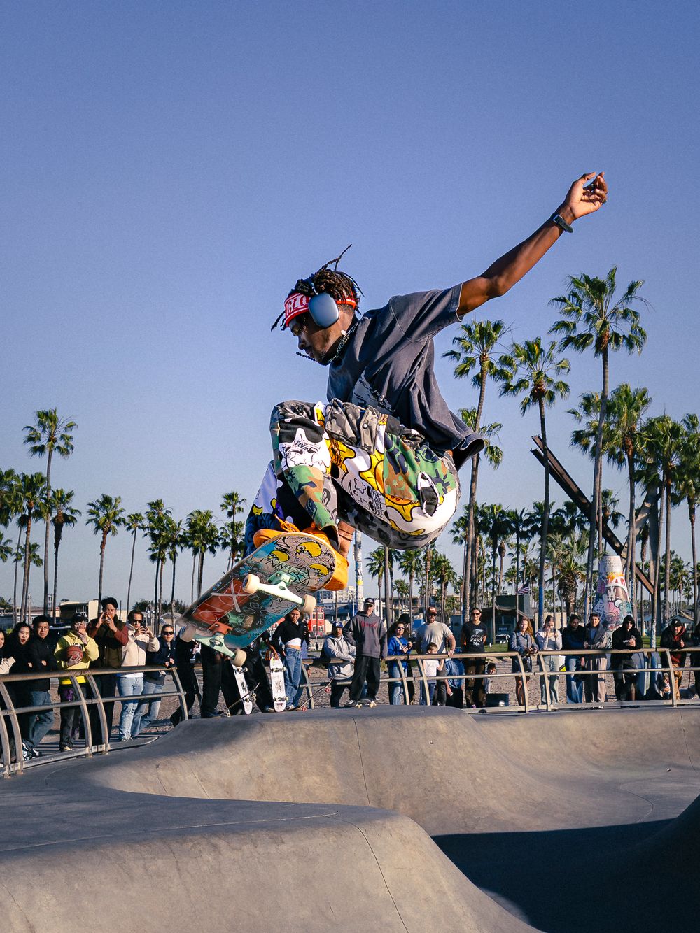 Skateboarder performing aerial trick at Venice Beach skate park with palm trees and crowd in background.