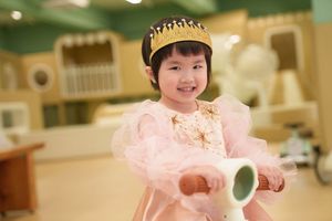 A smiling young girl wearing a pink princess dress and gold crown poses happily indoors.