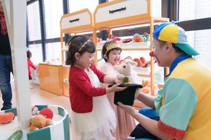 Children playing at a colorful supermarket roleplay station with a clown entertainer in a bright indoor play area.