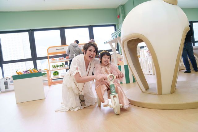 A woman and young girl smile together at an indoor children's play area with colorful toys and a ride-on toy bike.