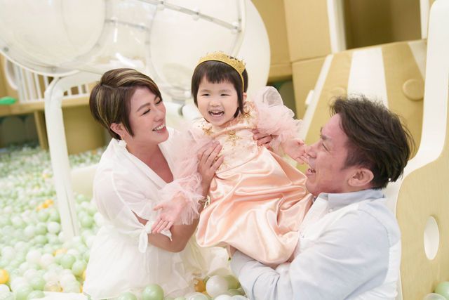 Happy family with parents lifting young girl in princess dress at colorful ball pit indoor playground.