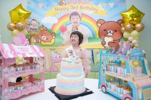 Young girl celebrates her 3rd birthday with a rainbow cake, Rilakkuma decorations, and colorful party backdrop.
