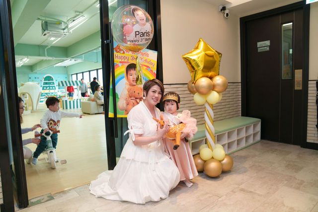 Woman in white dress poses with young girl in princess outfit beside gold balloons at a children's party venue.